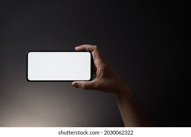 Colour image of man's hand holding black smartphone horizontally with blank white screen isolated on dark background with dramatic lighting and copy space to side