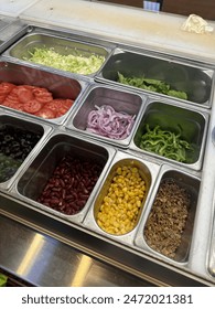 A colourful array of fresh salad toppings in a salad bar setup, including tomatoes, onions, and beans, suggests a healthy food option