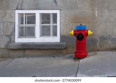 A colourful Class C hydrant painted red with yellow, black and blue caps. The fireplug is a dry-barrel underground hydrant. The hydrant is on a sidewalk in front of a brick building with a small glass window. 