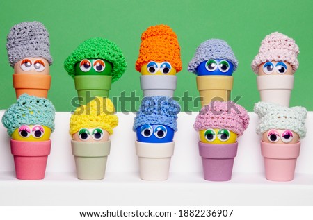 Colourful Easter eggs, made of recycled plastic, with wobbly eyes and colourful knitted hats, aligned horizontally and sitting in  bamboo egg holders against an apple green background. 