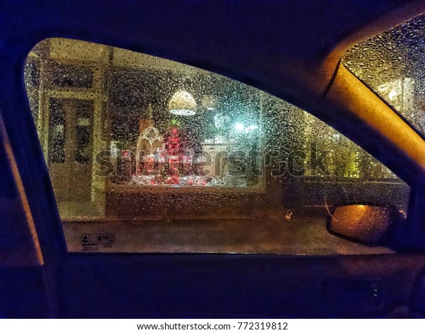 Colourful illuminated shop window display at\
night seen through car side window covered by raindrops. Abstract\
view of lit store window installation in wet dark night from\
automobile interior.