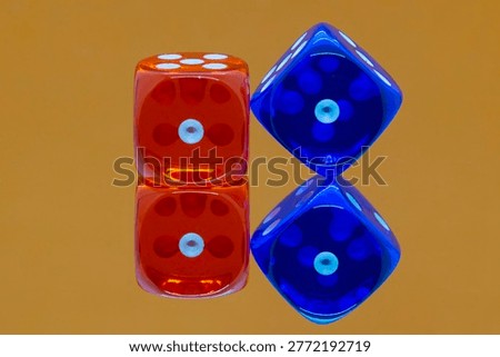 A colourful macro closeup image of 2 translucent dice reflected in a mirror against a bright orange background. The orange dice on the left shows a single dot and sits squarely, where the blue dice on the right sits on an edge in a diamond shape, also showing a single dot.