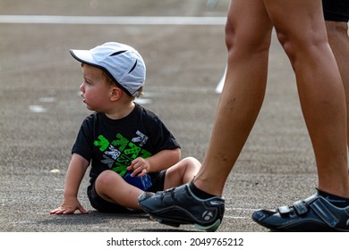 Columbus, OH USA 08-07-2021: A Tired Blond Toddler Boy Sitting On The Ground At A Parking Lot During An Event As His Parents Are Seen Besides Him. He Is Looking Away As He Touches Ground