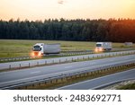 A column of truck tractors with semi-trailers and headlights on are transporting cargo along the highway against the backdrop of sunset and summer. Concept of working as a truck driver, remuneration