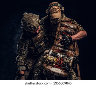 Combat Conflict, Special Mission, Retreat. Military Medic Rescues His Wounded Teammate Carrying Him Off The Battlefield. Studio Photo Against A Dark Wall