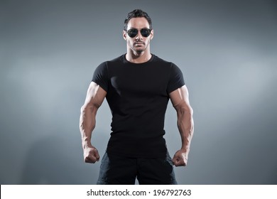 Combat muscled action hero man wearing black t-shirt with pants and sunglasses. Studio shot against grey.