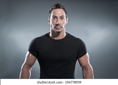 Combat Muscled Fitness Man Wearing Black Shirt And Pants. Studio Shot Against Grey.