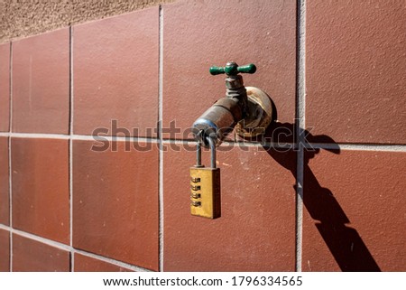  A combination lock on the end of an outdoor water faucet located on a red tiled wall . The image can be a metaphor for drought or water conservation.