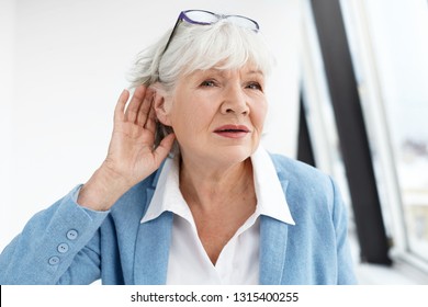 Come Again Please? Attractive Mature Woman Wearing Blue Jacket And Spectacles On Her Head Eavesdropping. Stylish Retired Female In Elegant Clothes Holding Hand At Her Ear, Suffering From Hearing Loss