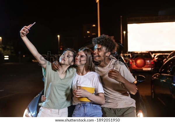 Come here\
and have fun. Cheerful diverse young friends smiling while taking a\
selfie together, standing in front of a big screen at open air\
cinema. Friendship, outdoor cinema\
concept