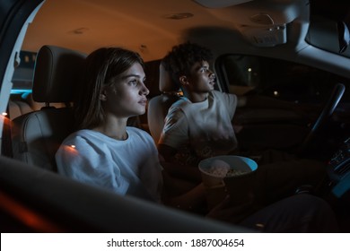 Come here and watch the movie. Diverse young couple having a romantic date. Young people holding popcorn and watching a movie at drive in cinema from the front seats of the car. Entertainment concept