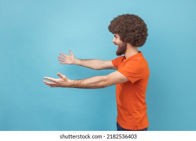 Come Into My Arms. Side View Of Satisfied Man With Afro Hairstyle Wearing Orange T-shirt Reaching Out To Camera, Stretching Arms To Hug You. Indoor Studio Shot Isolated On Blue Background.