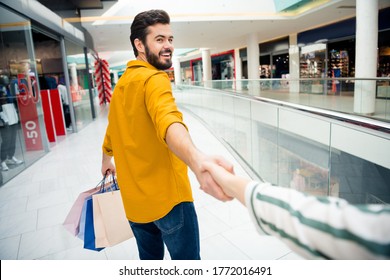 Come With Me. Photo Of Cheerful Handsome Guy Leading Girlfriend To Secret Surprise Place Carry Many Bags Walk Shopping Center Toothy Smiling Shopper Wear Casual Outfit Indoors