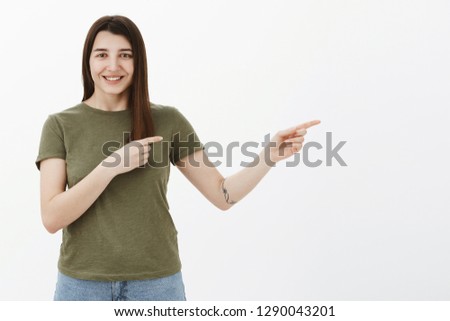 Come on I show you. Cheerful bright and happy young ordinary woman with tattoo and brown hair smiling delighted and friendly at camera as pointing right, indicating at cool place, leading way