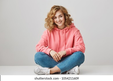 Come and sit with me. Portrait of cheerful relaxed young woman with curly hair in trendy clothes sitting on floor with crossed legs, smiling broadly while camping with friends in mountains - Shutterstock ID 1054211609