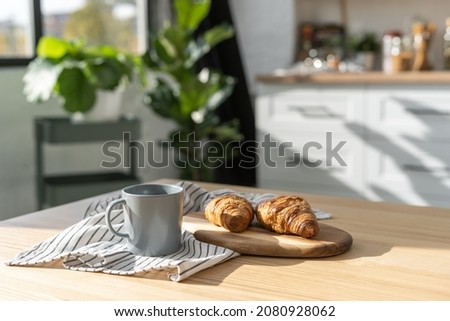 Comfort environment with tea and croissants in contemporary kitchen interior. Morning routine. Homemade baking concept. Modern white furniture and plants on background