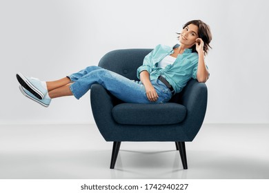 Comfort, People And Furniture Concept - Portrait Of Happy Smiling Young Woman In Turquoise Shirt And Jeans Sitting In Modern Armchair Over Grey Background