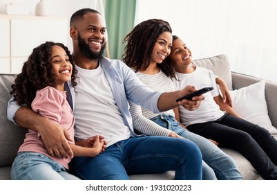 Comfort And Relax. Portrait Of Happy Loving Black Family Of Four People Watching Tv, Smiling Parents Embracing Their Two Children Sitting On Couch In The Living Room, Dad Holding Remote Control