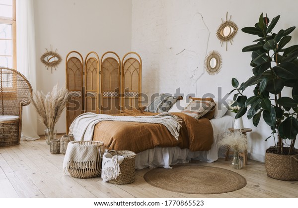 Comfortable
apartment in bohemian style interior with hygge bedroom, pillow and
bedspread on bed, bamboo dressing screen, home decor, dry plants in
vase, wicker basket, houseplant on
floor