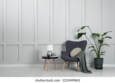 Comfortable armchair, table with lamp and houseplant near light grey wall in room, space for text. Interior design - Shutterstock ID 2219499841