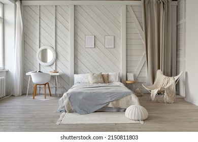 Comfortable bed with blanket and cushions placed on rug at wall with decorative frames in stylish bedroom with table and armchair in pastel colors