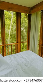 Comfortable bed inside a rustic wooden bedroom featuring large windows that overlook a lush green tropical forest jungle scene