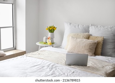 Comfortable bed with laptop, vase with tulips and Easter decor on table near light wall