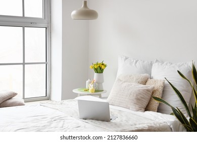 Comfortable bed with modern laptop, vase with tulips and Easter decor on table near light wall
