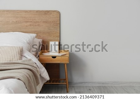 Comfortable bed and nightstand with blank frame near light wall