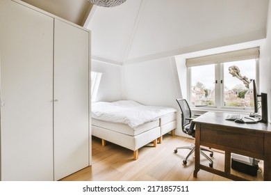 Comfortable Bed Opposite A Computer Desk With An Armchair, In An Attic Bedroom In A Modern Minimalist Style With Windows