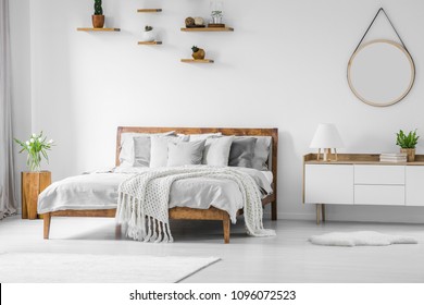 Comfortable, big, wooden bed with linen, pillows and blanket, nightstand beside and round mirror hanging on a white wall in a bright bedroom interior. Real photo.