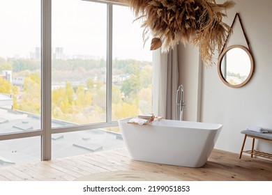 Comfortable bright bathroom with a boho-chic interior design, a free-standing white bath against the background of a panoramic window. hanging decor from dry grass. Soft selective focus.