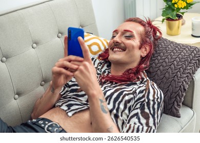Comfortable non-binary influencer with pink hair and mustache is using smartphone and smiling while lying on sofa, enjoying social media and online community