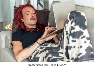 Comfortable non-binary person with red hair and painted eyebrows relaxing on the sofa at home, using a laptop and a tablet, enjoying modern technology and celebrating body positivity