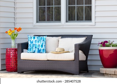 Comfortable Outdoor Living Area On A Brick Patio With A Deep Seating Settee And Cushions Flanked By Red Ceramic Pedestal Table And Flowerpot With Spring Flowers And A Straw Sunhat