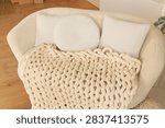 A comfortable seating area features a soft large blanket draped over a couch. Two small pillows sit beside a round pillow on the couch. The wooden floor adds warmth to the room.