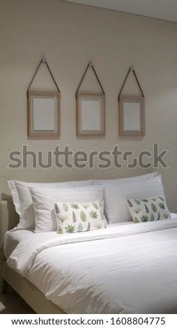 Comfortable throw cushion on the white pillow s and white bedsheet with three hanging picture frames in the bedroom                      