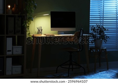 Comfortable workplace with computer and lamp on wooden desk at home office in evening