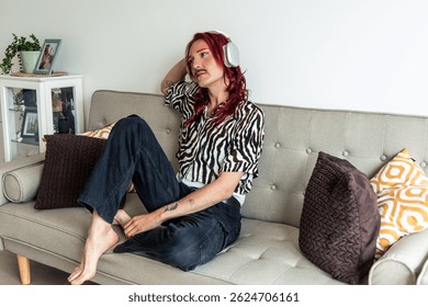 Comfortable young non-binary person with red hair and tattoos wearing headphones is sitting on a sofa at home listening to music, enjoying their free time in a body positive and inclusive way