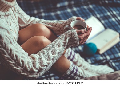 Comforting cozy woman in knitted winter socks and white warm sweater sitting on a plaid blanket and warming up with a cup of hot cocoa at home in winter time. Cozy time and winter drinks 