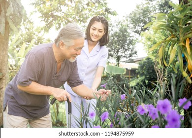 Comforting Hand. Young Nurse Holding Old Man's Shoulder In Outdoor Garden Looking At Flower. Senior Care, Care Taker And Senior Retirement Home Service Concept.