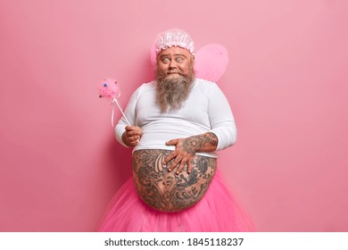 Comic Bearded Adult Man With Fat Belly Holds Magic Wand Acts Out Fairy Has Innocent Look Entertains His Children Has Long Thick Beard Models Against Pink Background. Costume Carnival Concept