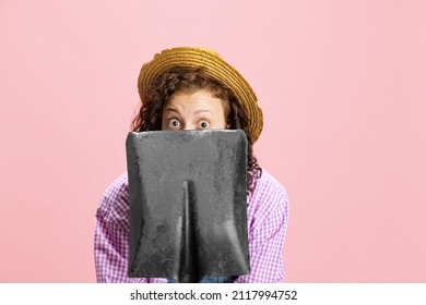 Comic Portrait Of Young Scared Girl, Gardener In Work Uniform And Straw Hat Hiding Behind Shovel Isolated On Pink Background. Concept Of Job, Emotions, Agronomy. Funny Meme Emotions. Copy Space For Ad