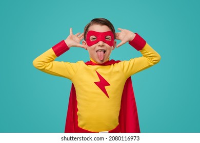 Comic preteen superhero in vivid costume and mask showing tongue and grimacing while looking at camera and having fun on blue background in studio
