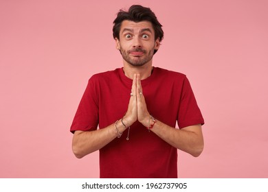 Comical excited young man with stubble in red tshirt making funny face and keeps hands in praying position over pink background