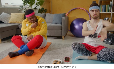 Comical Scene. Black Clumsy Ridiculous Guy Eating Cookies Taking Smartphone Pictures Of His Concentrated Funny Friend Meditating. Yoga At Home. Sports Parody.