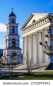A commanding view captures two of Chisinau's most significant religious architectural elements side by side against a deep blue winter sky. On the right, the grand portico of the Nativity Cathedral dominates the frame with a row of tall, fluted neoclassical columns supporting a large triangular pediment with clean, elegant proportions reminiscent of ancient Greek temple architecture. The cream-colored stone surface is bathed in bright sunlight, emphasizing the verticality and grandeur of the colonnade. Broad stone steps lead up to the cathedral entrance, and a decorative cast-iron street lamp with a spherical globe is mounted at the corner. Several pigeons are perched along the entablature frieze. To the left, the tiered bell tower rises independently against the sky, its four ascending square levels topped by a dark dome and a prominent Christian cross. Bronze bells are visible within the open upper belfry. A small rotunda structure is partially visible at the tower's base. In the foreground, a patch of green grass with traces of lingering snow surrounds a small bare-branched sapling. Pine tree branches frame the upper right corner, adding organic texture. The overall composition presents a powerful juxtaposition of the cathedral's horizontal classical breadth alongside the tower's soaring vertical thrust, conveying deep spiritual heritage and architectural magnificence.