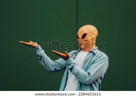 Commercial advice image portrait with green wall background to copywrite and write text and man in showing posture wearing alien ufo mask in casual clothes. Sales and discount adv concept