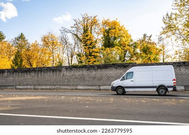 Commercial compact cargo small size white mini van delivering cargo to client running on the local road with autumn yellow trees and concrete protection fence on the side