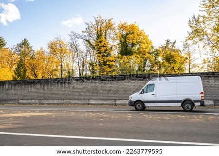 Commercial compact cargo small size white mini van delivering cargo to client running on the local road with autumn yellow trees and concrete protection fence on the side
